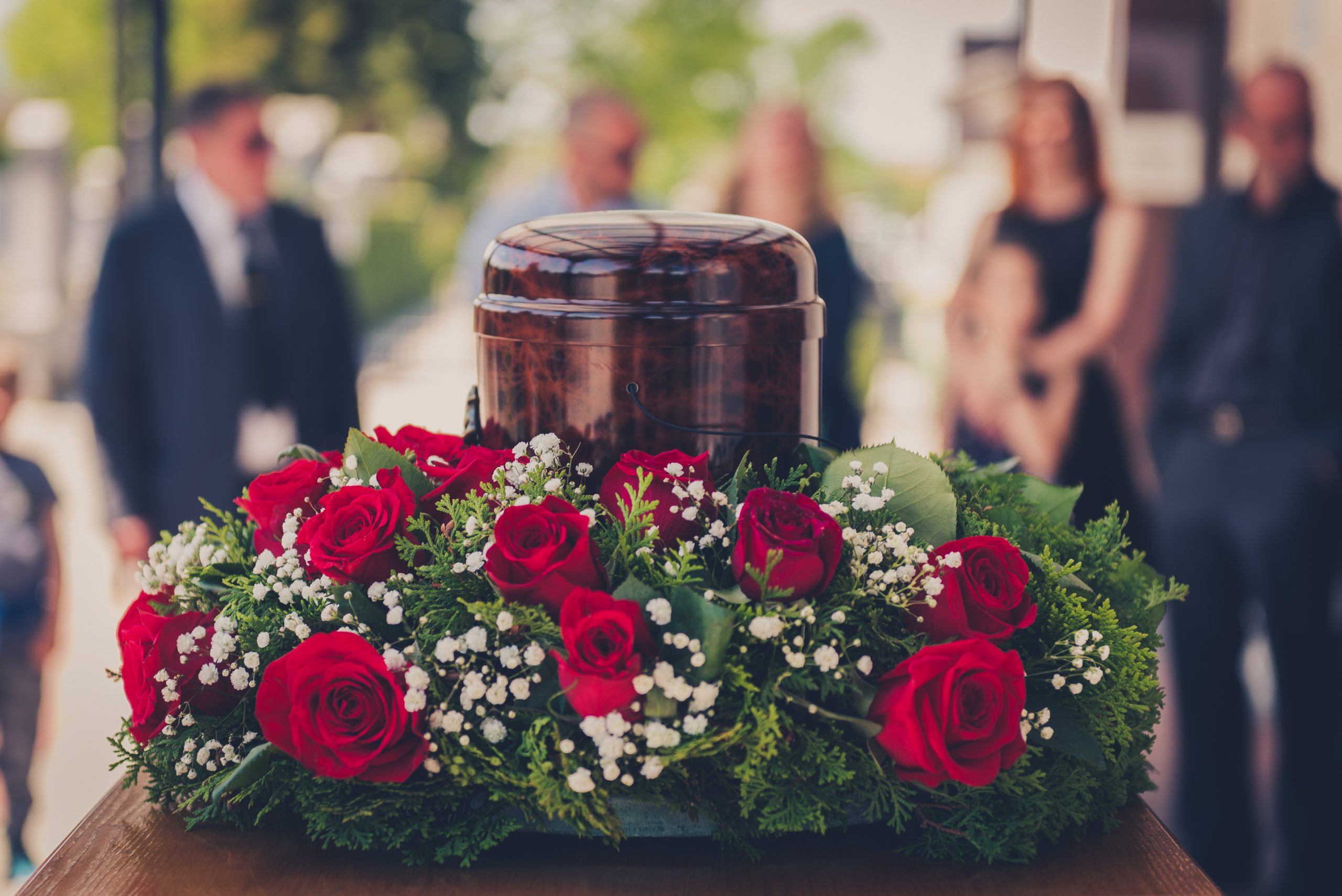 People attend a funeral with an urn in the foreground surrounded by a rose wreath